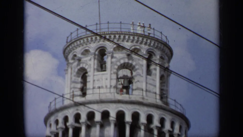 1962 Italy home-movie view of the tower belfry with visible bells, rooftop railing, blue sky, clouds, and crossing wires.