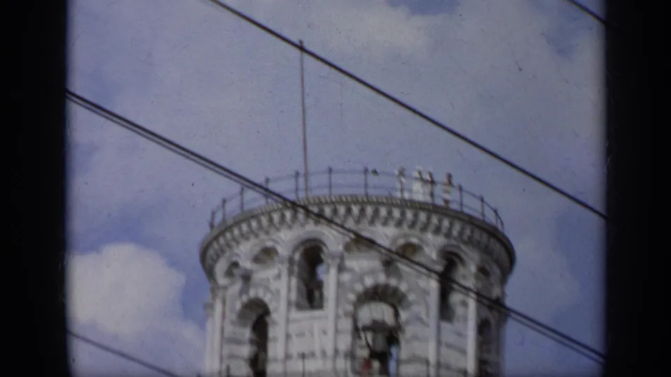 1962 Italy 8mm frame showing the Leaning Tower of Pisa belfry, bells, rooftop railing, blue sky, clouds, and diagonal wires.