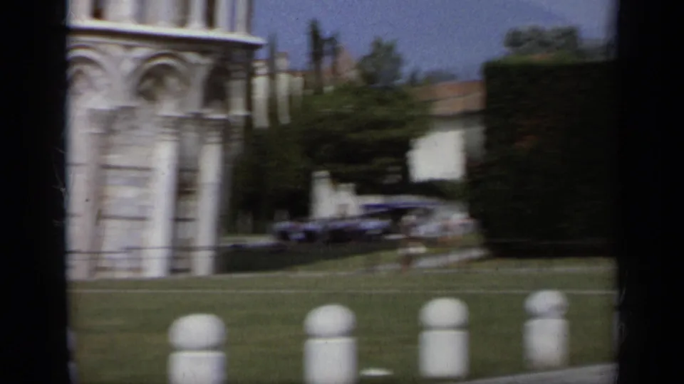 1962 8mm frame showing the Pisa tower area with lawn, nearby architecture, parked cars, hedges, and small distant people.