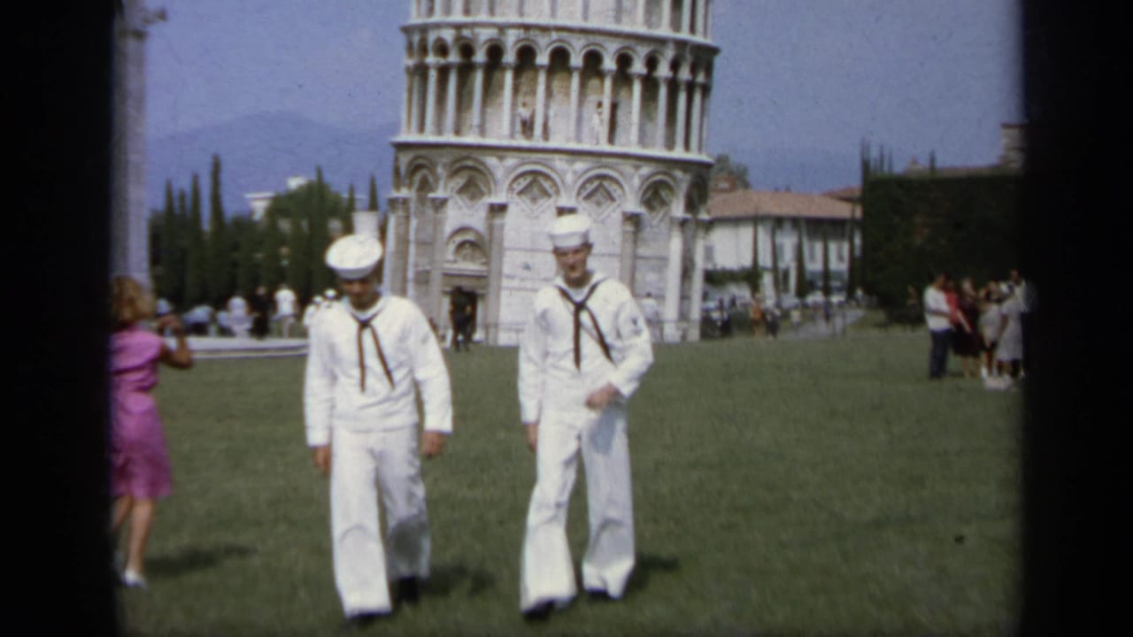 Two sailors in white uniforms approach the camera at Pisa's Leaning Tower while tourists stand behind them, 1962.