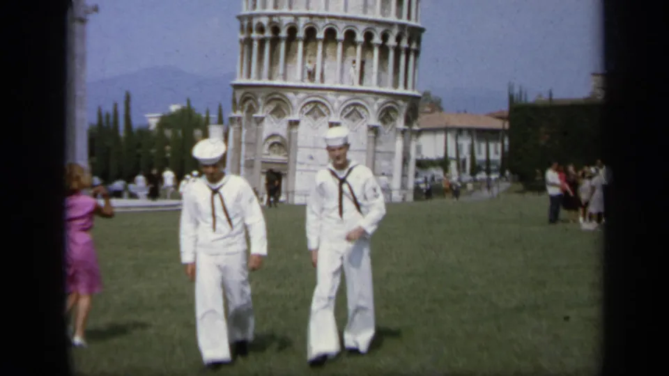 Two sailors in white uniforms approach the camera at Pisa's Leaning Tower while tourists stand behind them, 1962.