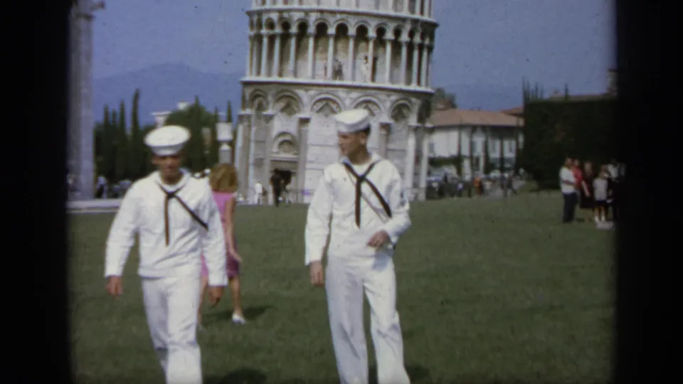 Two sailors in white uniforms pass close to the camera in Pisa as a woman in a pink dress and other tourists appear behind them.