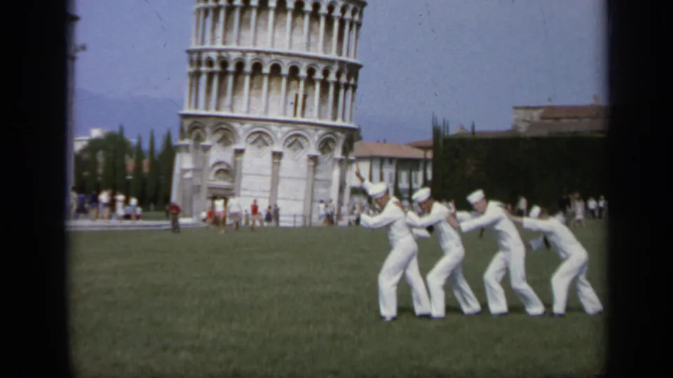 1962 Italy home-movie frame of four sailors posing on grass as if pushing near the Leaning Tower of Pisa, with tourists behind them.