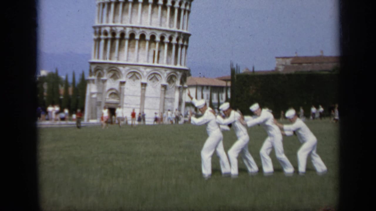 Sailors in white lean forward on grass with the Leaning Tower of Pisa behind them.