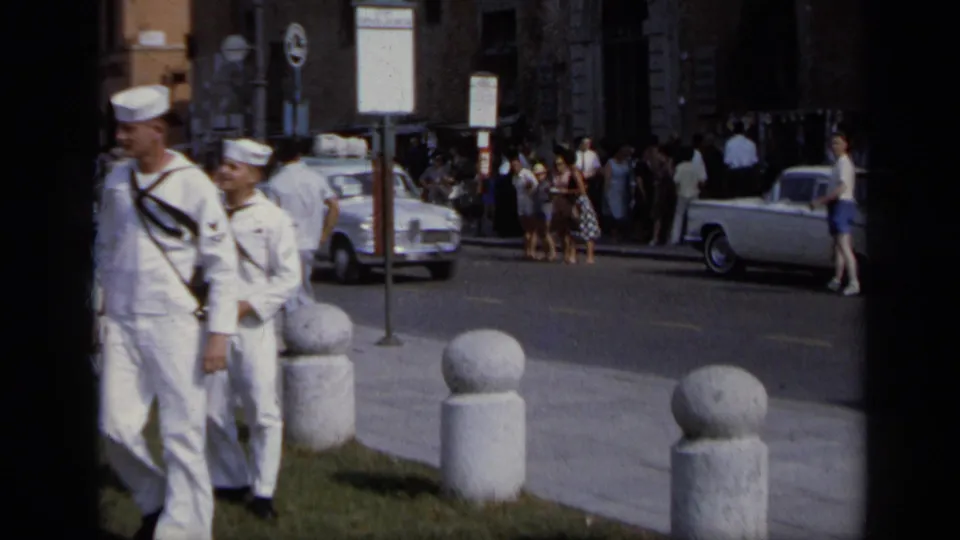 1962 Italy home-movie frame showing two U.S. Navy sailors walking near stone bollards, street signs, cars, and a crowd by buildings.