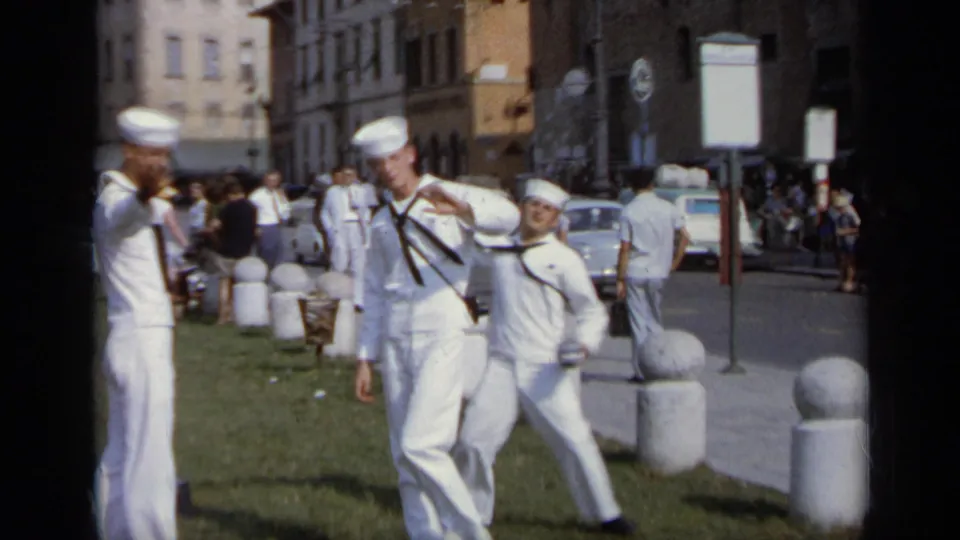 1962 Italy frame showing uniformed sailors clowning for the camera near stone bollards, with tourists and parked cars in the background.