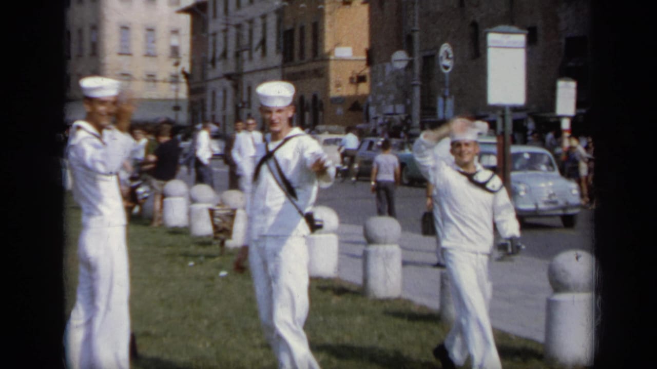Three sailors in white uniforms stand on grass beside a city sidewalk and road.