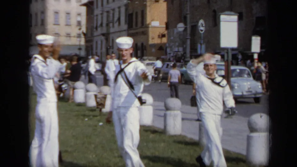 In 1962 Italy, three U.S. Navy sailors in white uniforms stand and wave near grass, bollards, parked cars, and pedestrians.