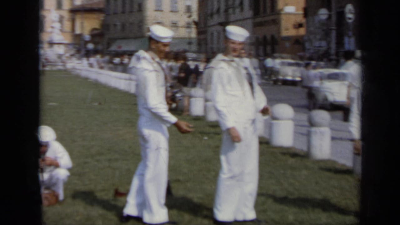 Two U.S. Navy sailors in white uniforms stand in a sunny Italian park area with a road, bollards, cars, and pedestrians behind them.
