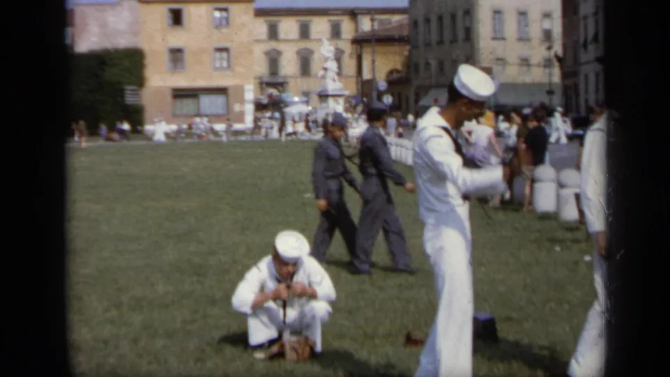 A sailor crouches to handle a camera or gear while another sailor stands nearby in a public park area in Italy.