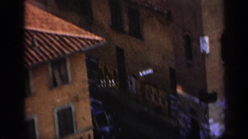 Handheld 1962 Italy travel frame showing terracotta rooflines, tall walls, shuttered windows, and a shadowed narrow passage.