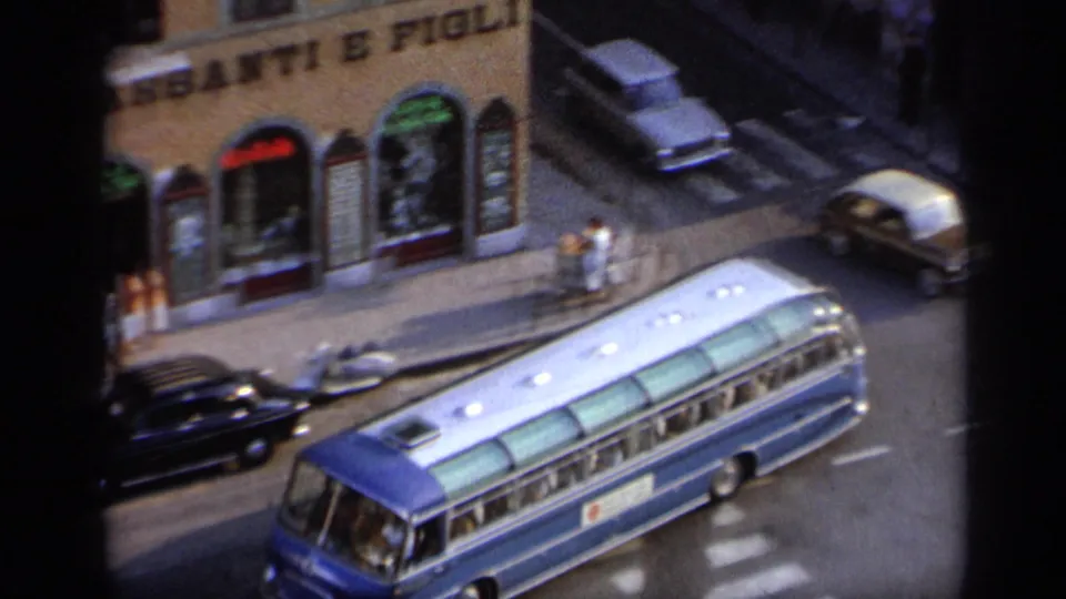1962 Italy street view from above with a blue coach bus, vintage cars, pedestrians, and storefront-style building fronts.