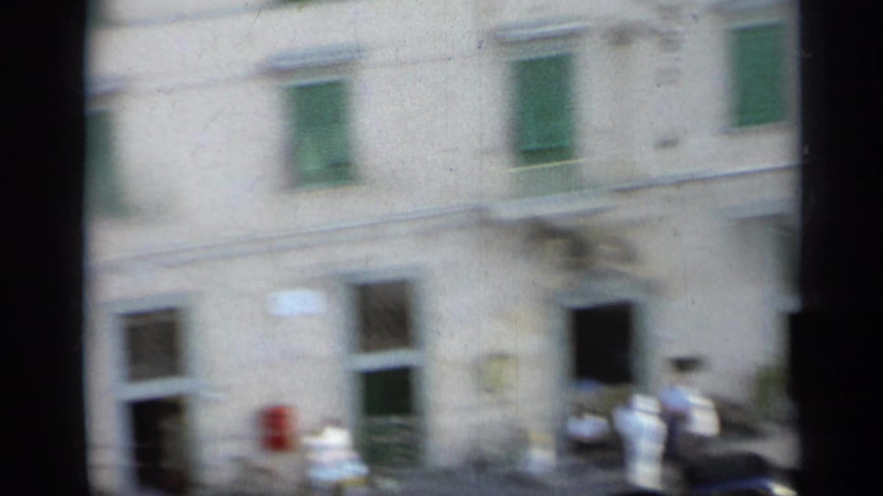 Soft, handheld 8mm view of an Italian building facade with green shutters and a few pedestrians near the street level.