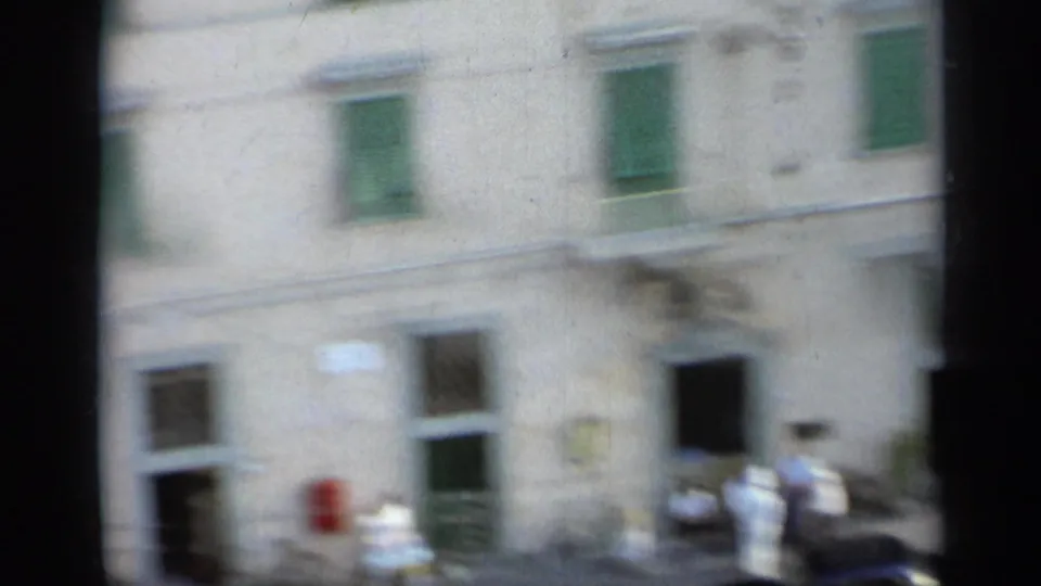 Soft, handheld 8mm view of an Italian building facade with green shutters and a few pedestrians near the street level.