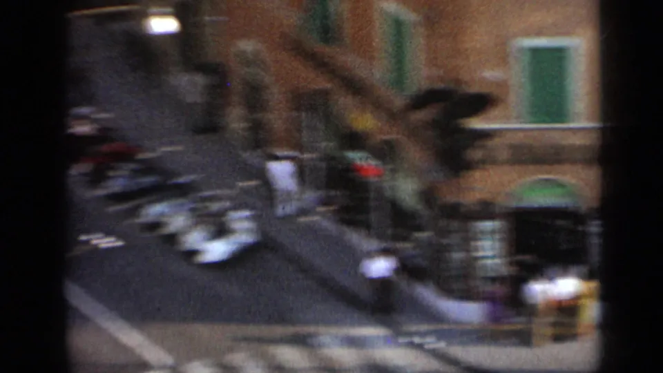 Handheld elevated view of a 1962 Italian street corner with vintage cars, pedestrians, arched storefronts, and green shutters.