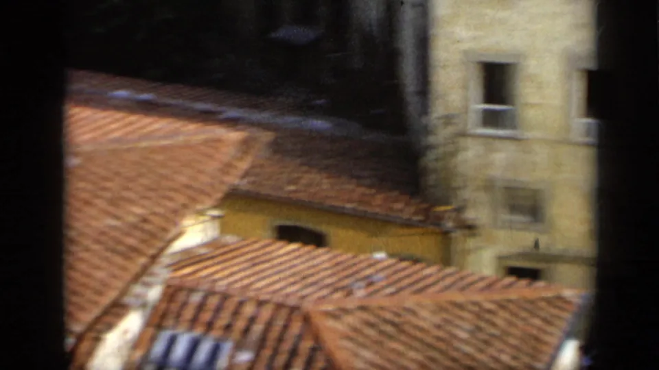 Elevated 8mm view in Italy showing terracotta roof tiles, pale building walls, windows, and a narrow urban courtyard area.