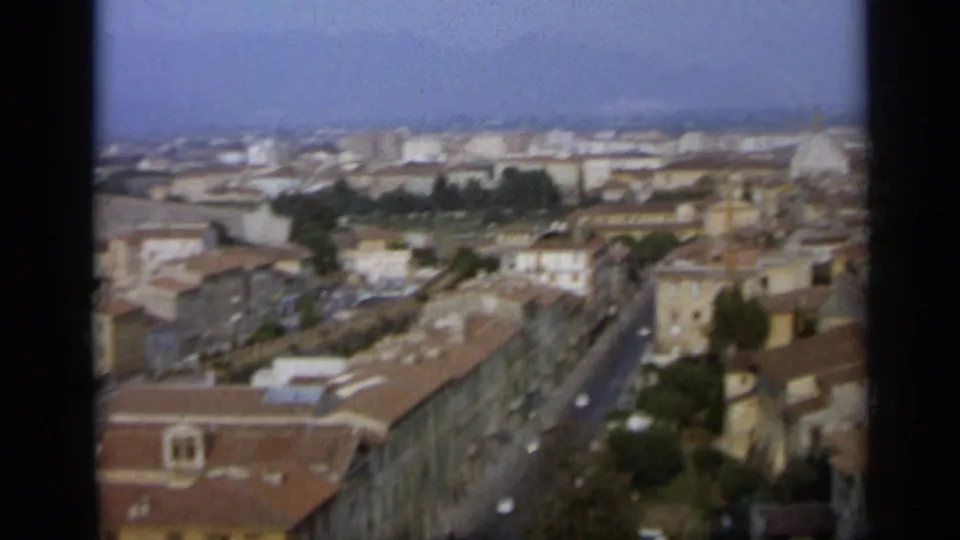 1962 Italian city panorama with a straight street, rows of tiled roofs, pale masonry buildings, and a dome in the distance.