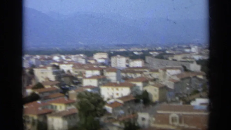 1962 8mm frame of an Italian urban district, with tiled roofs, light-colored buildings, trees, and distant hills softened by haze.