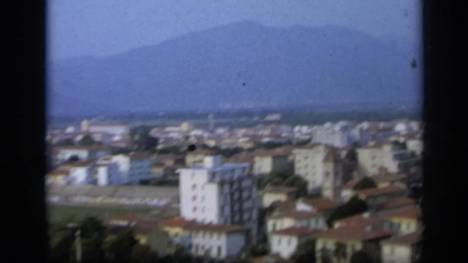 1962 Italian city view showing apartment blocks, terracotta roofs, open ground, and a hazy mountain backdrop.