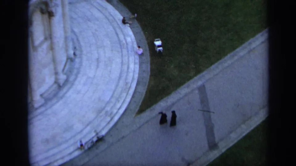 High-angle 1962 Assisi view with two people in dark clothing walking on a pale stone walkway beside the basilica and a grass area.