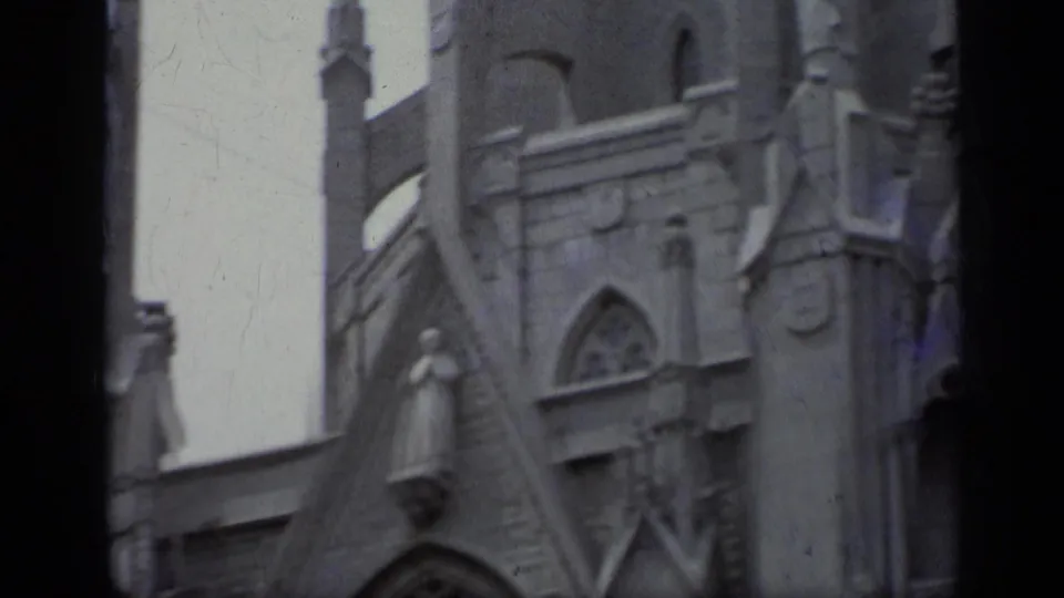 1962 Italy 8mm frame showing a pale stone church facade with Gothic-style arch, spire, sculptural ornament, and soft handheld framing.
