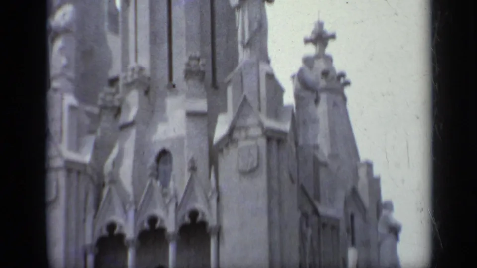 1962 Italy 8mm frame of an ornate church tower section with a cross, carved figures, pointed openings, and pale stone ornament.