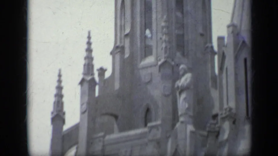 1962 Italy 8mm frame showing a visitor's close view of a stone church facade with a statue, spire, arched detail, and carved trim.