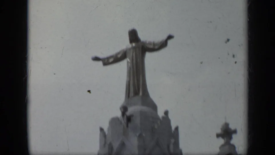 1962 Italy 8mm frame showing a robed Jesus statue with spread arms on top of a cathedral tower, seen from below.