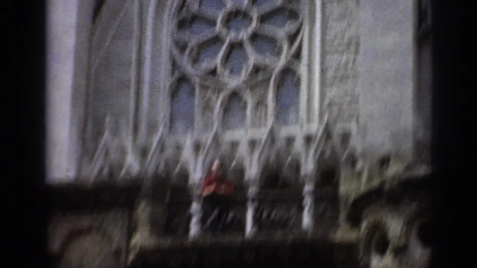 1962 Italy 8mm frame showing Gothic facade details, a rose-window form, balcony railing, and a distant person in red.