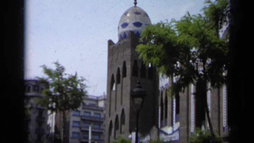 1962 Italy 8mm frame showing a church tower with arched windows and a blue-tiled dome near trees and urban buildings.