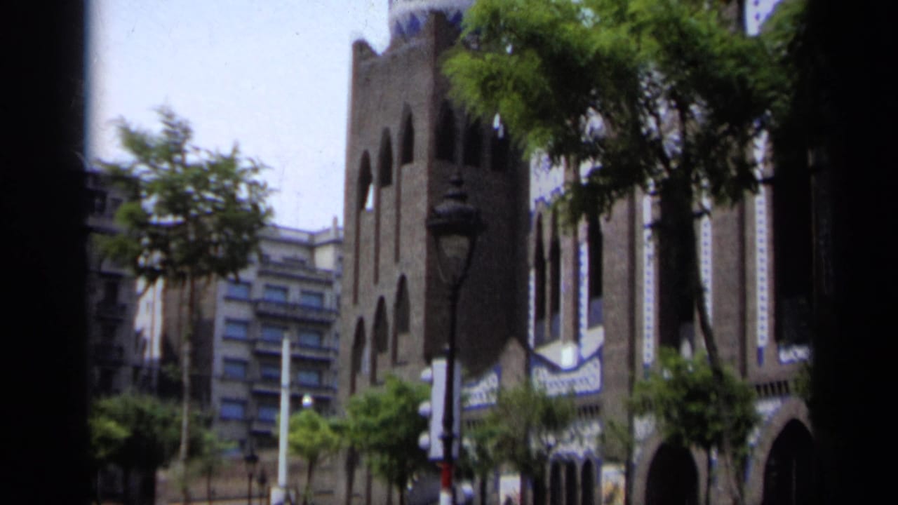 Gothic-style building facade with arched windows, trees, and street lamps across a sunlit street.