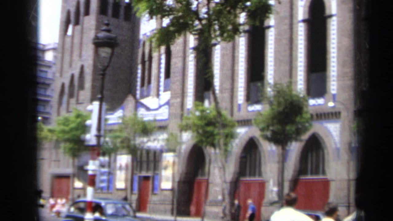 Facade with red doors, arched windows, trees, pedestrians, and a dark car near the curb.