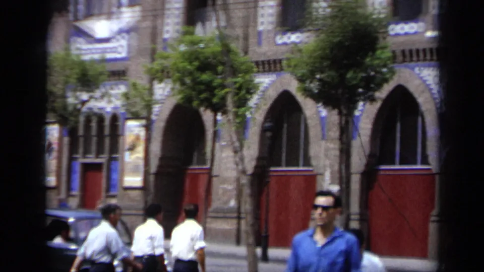 1962 Italy home-movie frame showing men on the sidewalk in front of a colorful arched facade with red doors and trees.