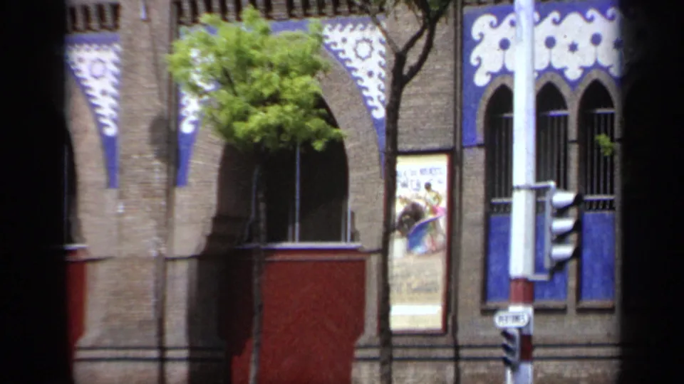 1962 Italy street frame with decorative blue and white trim, arched windows, a red door, tree branches, poster, and traffic signal.