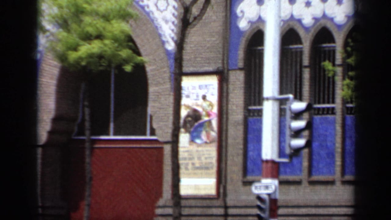 Decorated brick building with arched windows, blue panels, trees, traffic lights, and a bullfight poster.