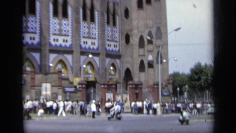 1962 Italy 8mm frame showing the decorated monument from across the street, with distant people and scooters at the base.