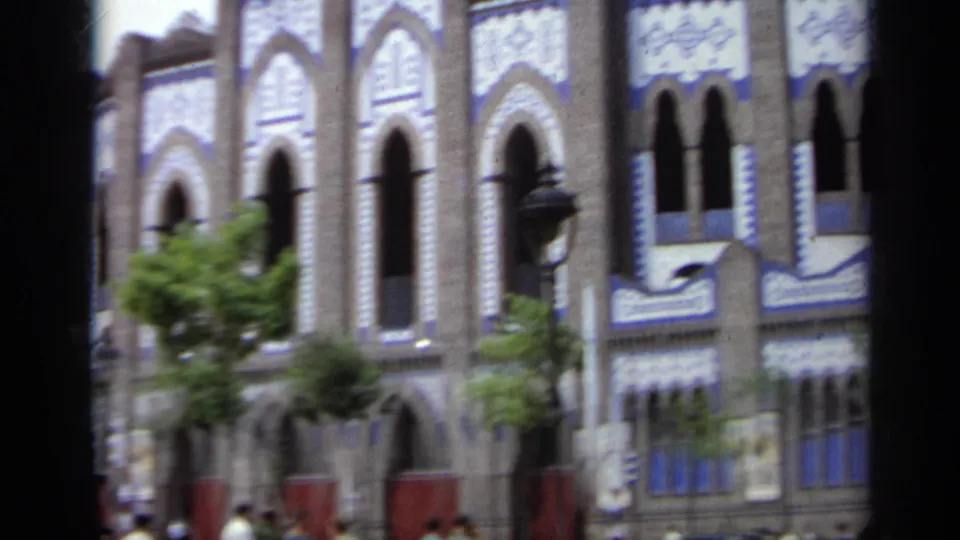 1962 Italy 8mm frame showing the stadium wall, repeated arched openings, blue trim, trees, and soft handheld motion blur.