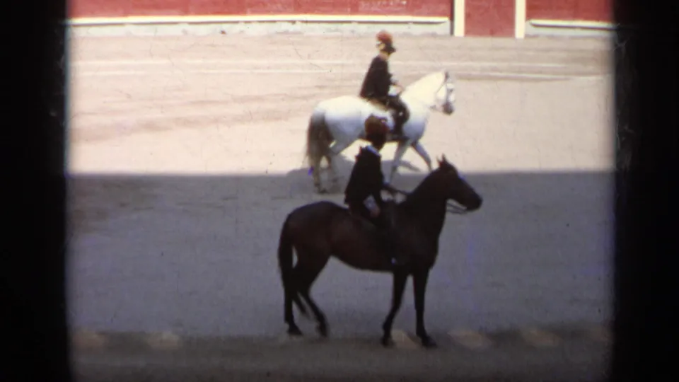 1962 Italy 8mm frame showing two distant riders on horseback moving across a sandy arena floor, one on a white horse and one on a dark horse.