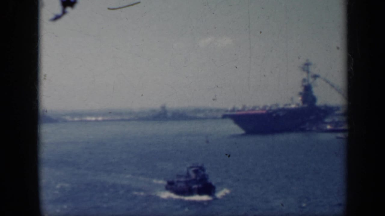 Small tugboat moves in front of a docked aircraft carrier in a harbor.