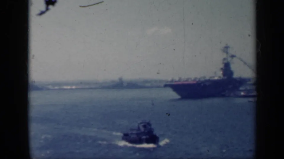Soft, blue-tinted 8mm view of a tugboat passing near a docked aircraft carrier in Italy, 1962.