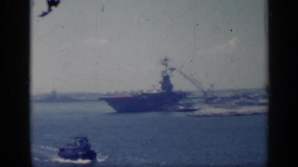 Grainy 1962 Italy harbor frame with a docked aircraft carrier and a small tugboat moving below it.