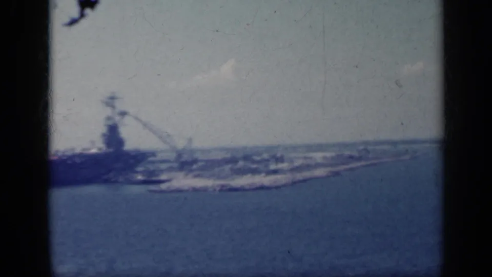 Blue-shifted 8mm frame from Italy in 1962 showing an aircraft carrier docked beside harbor structures.