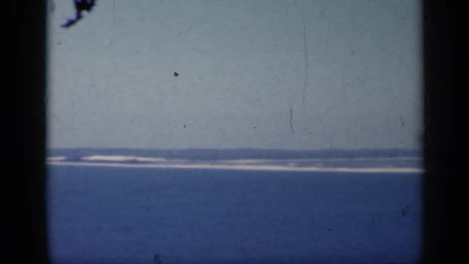 1962 Italy 8mm frame with a distant coastal strip, white surf, blue water, and soft handheld home-movie framing.