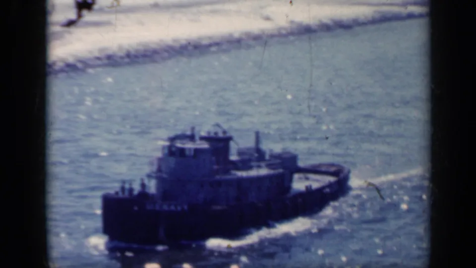 1962 Italy 8mm frame showing a dark brown tugboat crossing sunlit blue water with wake and coastline beyond.