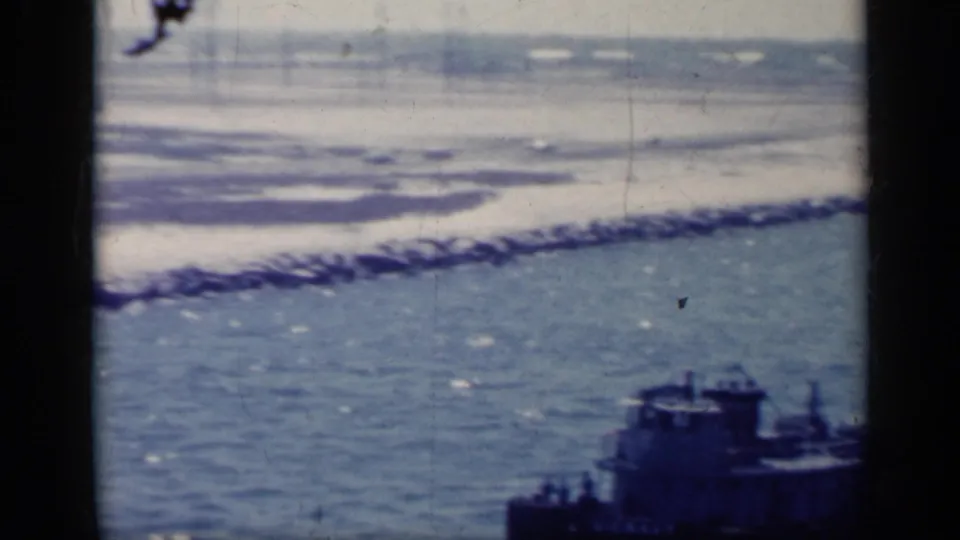 1962 Italy home-movie frame showing blue coastal water, a dark tugboat, rocky breakwater, and distant shoreline through dark foreground bars.