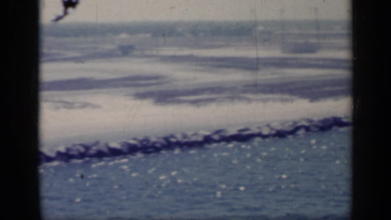 1962 Italy coastal view with low waves, a rocky breakwater, and faint horizon structures, framed by dark vertical edges.