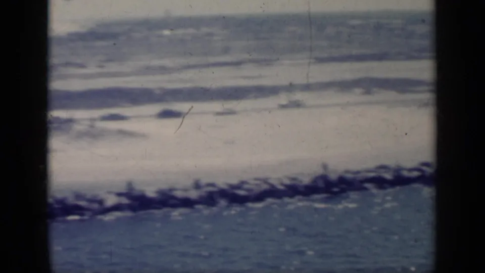 Faded 1962 Italy 8mm frame of coastal water, a rocky breakwater, and hazy distant land or structures seen through dark bars.