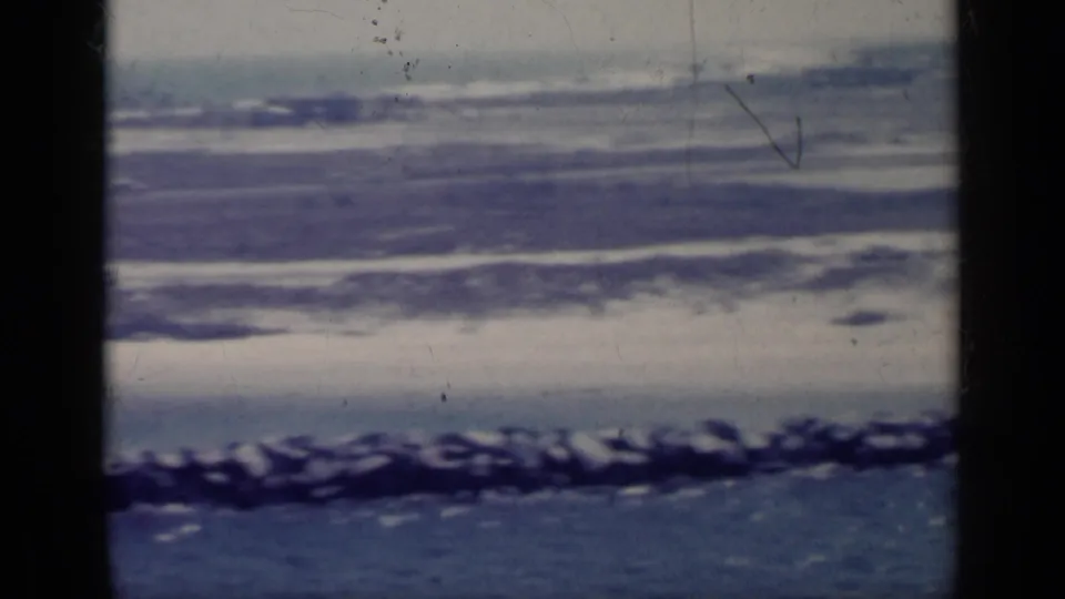 1962 Italy coastal home-movie frame with blue water, low waves, rocky shoreline, and hazy distant land, marked by visible film wear.