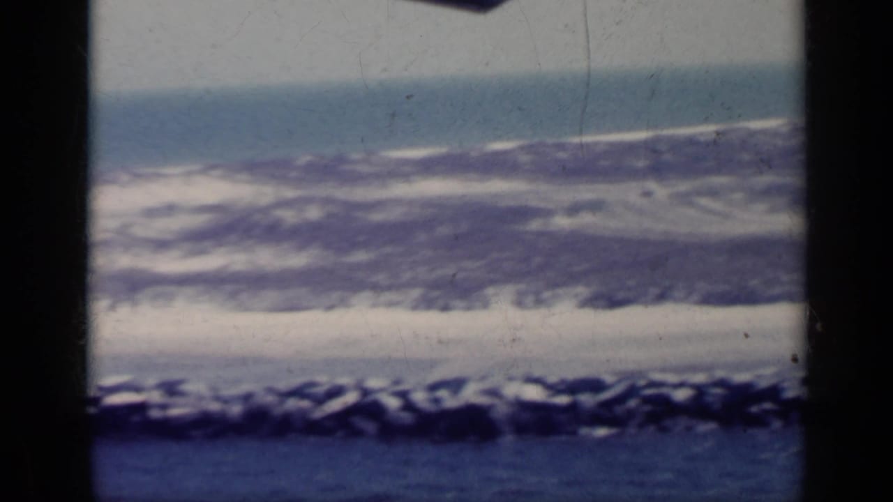 Low shoreline and pale surf beyond blue water, framed by dark vertical film edges.