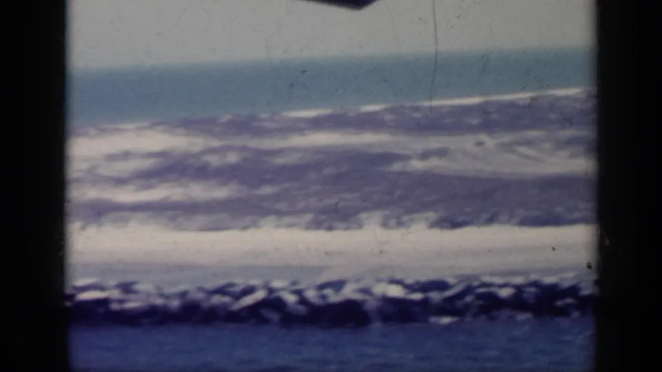 1962 Italy 8mm view of a pale coastal strip and surf beyond blue water, with dark gate edges and aged color shift.